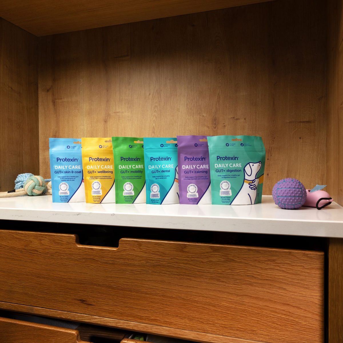 Five bags of Proteinix Daily Care dog supplements in different colors (blue, yellow, green, teal, and purple) lined up on a white countertop inside a wooden cabinet, with dog toys on either side.