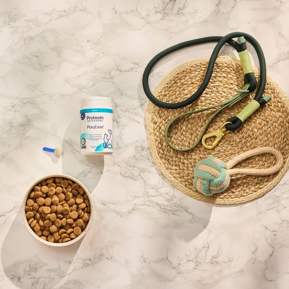A styled flat-lay of pet accessories and treats on a white marble surface, including a bowl of kibble, a supplement bottle, a green leash, and a woven mat with a toy.